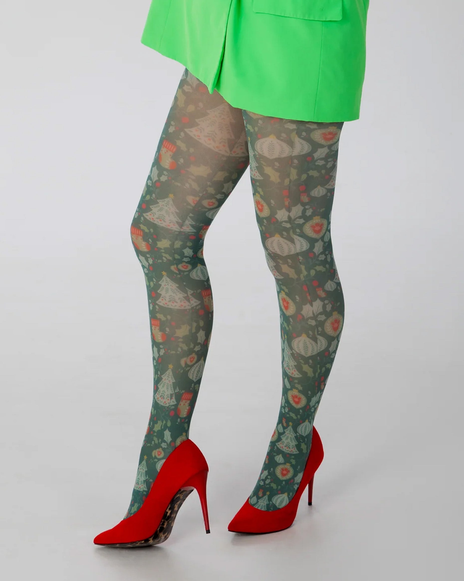 Pamela Mann Christmas Chaos Tights - White opaque tights with a Christmas themed print of Xmas trees, baubles, stockings & holly in shades of green and red. Worn with neon green blazer and red high heels.